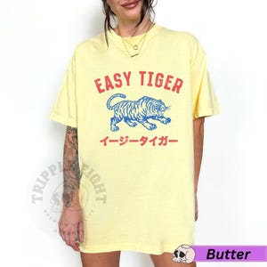May include: Pale yellow t-shirt featuring the red text "EASY TIGER" above a blue tiger illustration. Japanese text is below the tiger. The shirt has a loose fit.