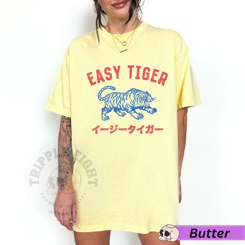May include: Pale yellow t-shirt featuring the red text "EASY TIGER" above a blue tiger illustration. Japanese text is below the tiger. The shirt has a loose fit.