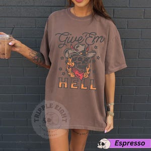 May include: Brown t-shirt featuring a skull wearing a cowboy hat and flames. The text "Give 'Em Hell" is printed on the shirt.