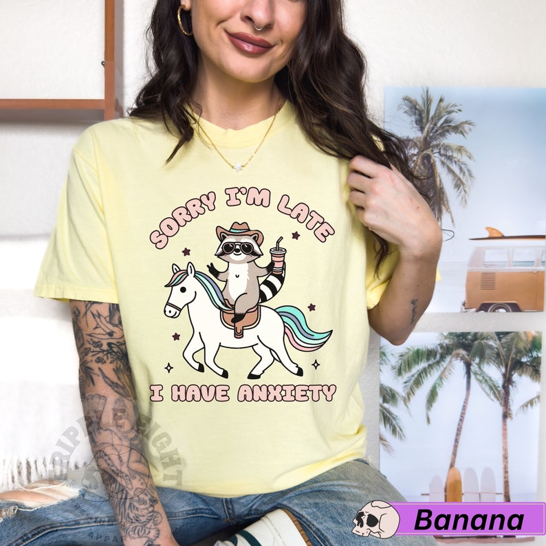 May include: Yellow t-shirt with a cartoon raccoon riding a unicorn. The text on the shirt reads "Sorry I'm Late I Have Anxiety".