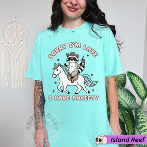 May include: A turquoise blue t-shirt with the text "Sorry I'm Late I Have Anxiety" in pink and white lettering. A cartoon raccoon wearing a cowboy hat is riding a white unicorn with a rainbow mane.
