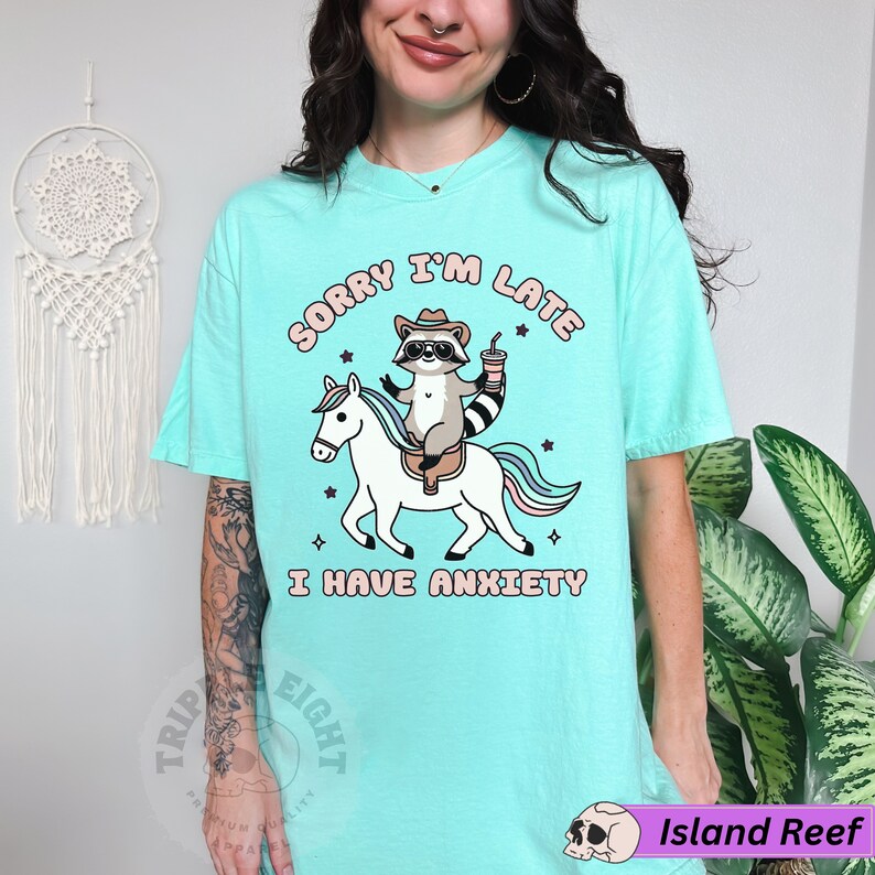 May include: A turquoise blue t-shirt with the text "Sorry I'm Late I Have Anxiety" in pink and white lettering. A cartoon raccoon wearing a cowboy hat is riding a white unicorn with a rainbow mane.