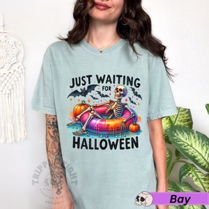 May include: A light blue t-shirt with the text "Just Waiting for Halloween" in black. A skeleton is sitting in a colorful inflatable ring with a pumpkin in each hand. The skeleton is wearing a black and white striped shirt and has a black and white striped hat.