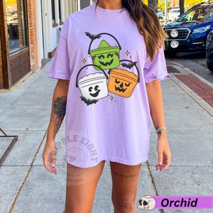 May include: A lavender t-shirt featuring a graphic of three Halloween buckets with different coloured lids and faces. The buckets are green, white and orange. The shirt has the words "Purple Eight" on the bottom left side.