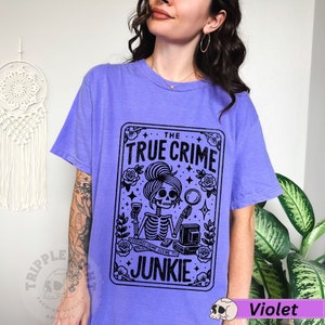 Puede incluir: Una camiseta morada con un gr&aacute;fico negro de una mujer esqueleto sosteniendo una lupa y una copa de vino. El gr&aacute;fico est&aacute; en un marco tipo tarjeta con el texto "The True Crime Junkie".