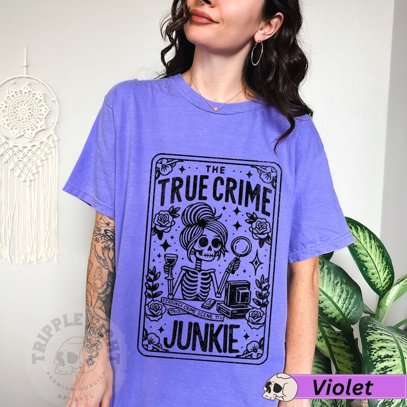 Puede incluir: Una camiseta morada con un gr&aacute;fico negro de una mujer esqueleto sosteniendo una lupa y una copa de vino. El gr&aacute;fico est&aacute; en un marco tipo tarjeta con el texto "The True Crime Junkie".