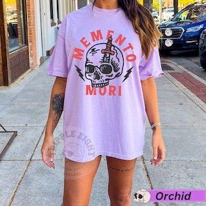 Puede incluir: Una camiseta de color lavanda con un gr&aacute;fico de una calavera con una daga que la atraviesa. El texto "Memento Mori" est&aacute; impreso en rojo debajo de la calavera.