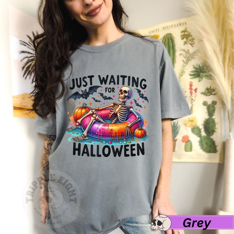 May include: A gray t-shirt with the text "Just Waiting for Halloween" and a skeleton in a donut pool float. The skeleton is wearing a pink and orange float with a pink and orange donut.