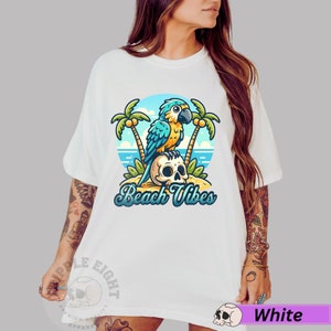 May include: White t-shirt with a blue and yellow parrot sitting on a skull with palm trees and the text "Beach Vibes".