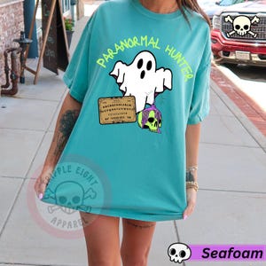 May include: Seafoam green t-shirt with the words "PARANORMAL HUNTER" in neon green. The shirt features a white ghost holding a Ouija board and a green skull with purple hair. The shirt is a comfortable, oversized fit.