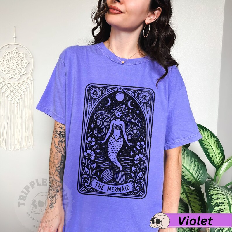 May include: A purple t-shirt featuring a black and white illustration of a mermaid in a tarot card style design. The text "The Mermaid" is printed below the illustration.