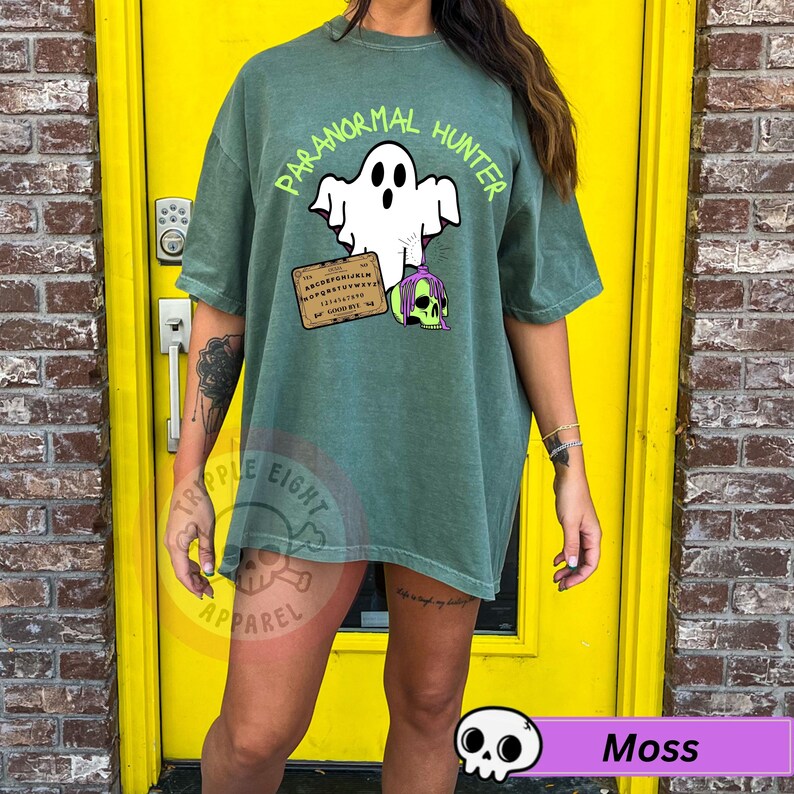 May include: Moss green t-shirt with the text "PARANORMAL HUNTER" above a white ghost graphic. Below the ghost is a planchette and a purple and green skull. The shirt is part of the Tripple Eight Apparel collection.