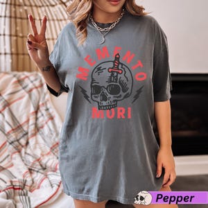 Puede incluir: Una camiseta gris con un gr&aacute;fico de calavera y el texto "Memento Mori" en rojo.