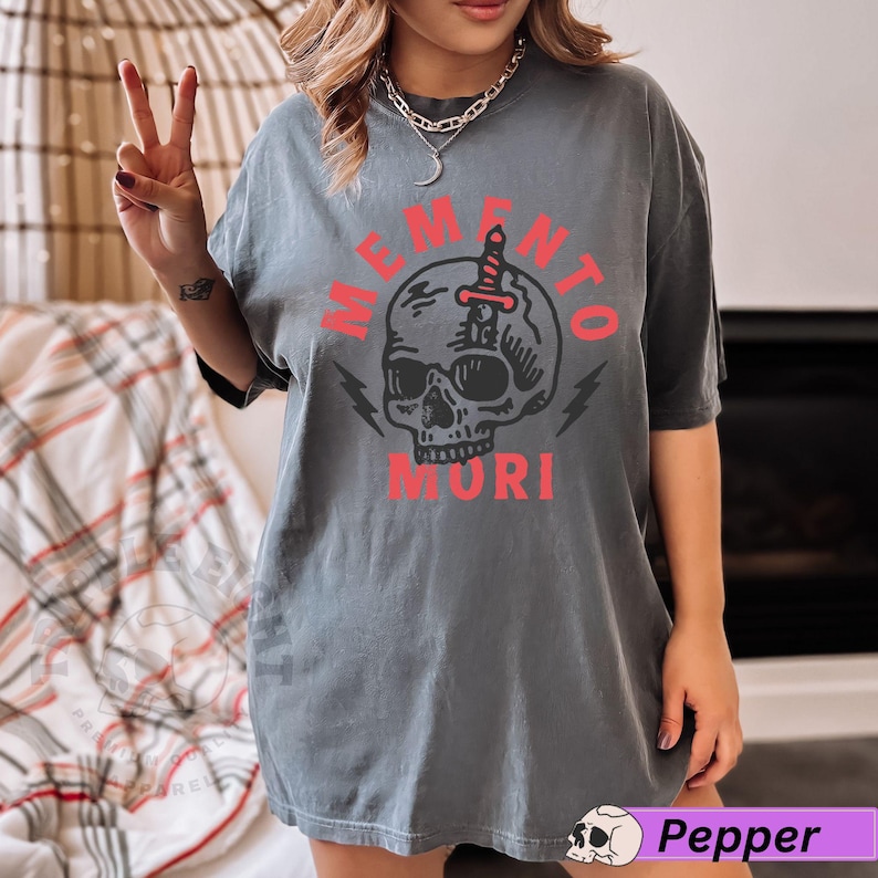 Puede incluir: Una camiseta gris con un gr&aacute;fico de calavera y el texto "Memento Mori" en rojo.