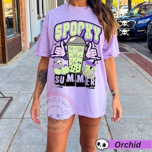 Puede incluir: Camiseta lila claro con un gr&aacute;fico de "Spooky Summer". El dise&ntilde;o incluye una mano de esqueleto sosteniendo una bebida, calaveras y calabazas. La camiseta tiene un corte holgado y es de estilo casual.