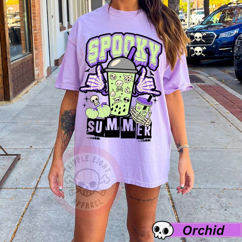 Puede incluir: Camiseta lila claro con un gr&aacute;fico de "Spooky Summer". El dise&ntilde;o incluye una mano de esqueleto sosteniendo una bebida, calaveras y calabazas. La camiseta tiene un corte holgado y es de estilo casual.