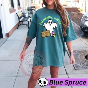 May include: Blue spruce t-shirt with a graphic of a ghost, a Ouija board, and a skull with purple hair. The text "PARANORMAL HUNTER" is above the ghost. The shirt also has the text "TRIPPLE EIGHT APPAREL" and "Blue Spruce".