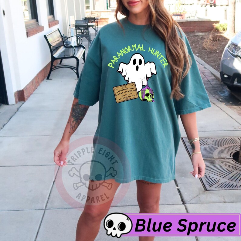 May include: Blue spruce t-shirt with a graphic of a ghost, a Ouija board, and a skull with purple hair. The text "PARANORMAL HUNTER" is above the ghost. The shirt also has the text "TRIPPLE EIGHT APPAREL" and "Blue Spruce".