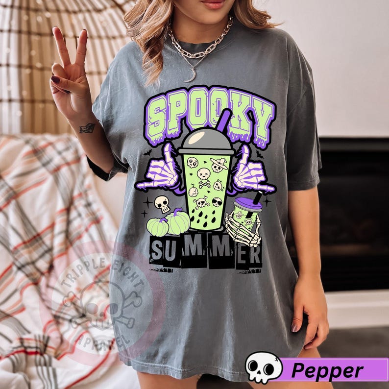 Puede incluir: Camiseta gris con un dise&ntilde;o de Halloween. El dise&ntilde;o presenta la palabra "Spooky" en verde y morado, junto con una bebida boba con calaveras y manos de esqueleto. La palabra "Summer" est&aacute; debajo.