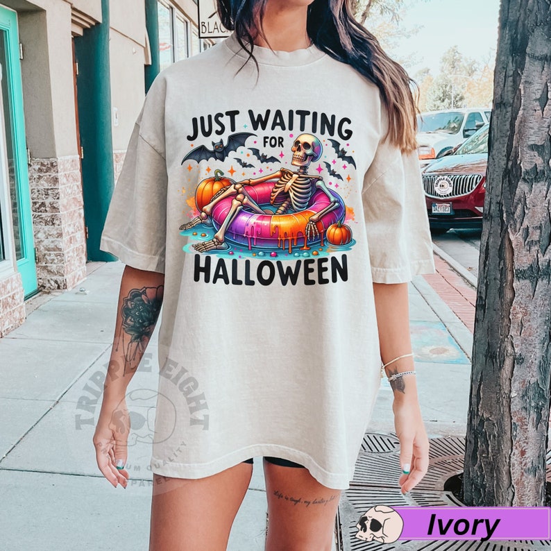 May include: A light beige t-shirt with a skeleton wearing a colorful inflatable ring and the text "Just Waiting for Halloween".