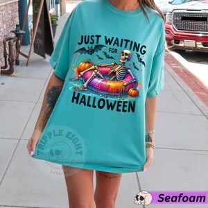 May include: A turquoise blue t-shirt with the text "Just Waiting for Halloween" and a skeleton wearing a colorful inflatable ring. The skeleton is surrounded by bats and pumpkins.