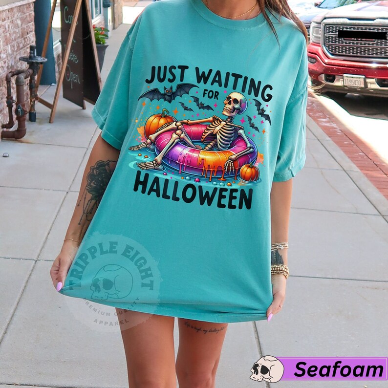 May include: A turquoise blue t-shirt with the text "Just Waiting for Halloween" and a skeleton wearing a colorful inflatable ring. The skeleton is surrounded by bats and pumpkins.