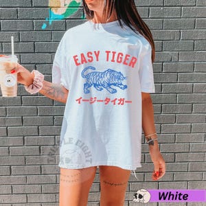 May include: White t-shirt featuring the text "EASY TIGER" in red, above a blue tiger illustration. Japanese text is below the graphic. The shirt has a relaxed, oversized fit.