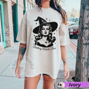 May include: A woman wearing a light beige oversized t-shirt with a black and white illustration of a witch with the text "Resting Witch Face".