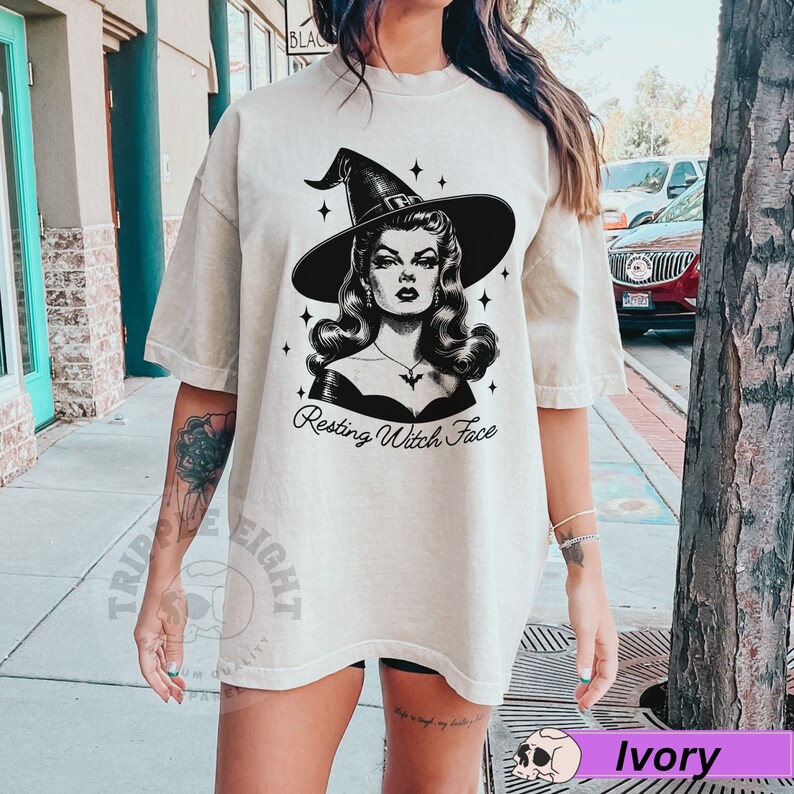 May include: A woman wearing a light beige oversized t-shirt with a black and white illustration of a witch with the text "Resting Witch Face".