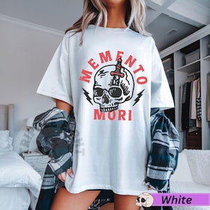 Puede incluir: Camiseta blanca con un gr&aacute;fico de calavera y el texto "Memento Mori" en rojo.