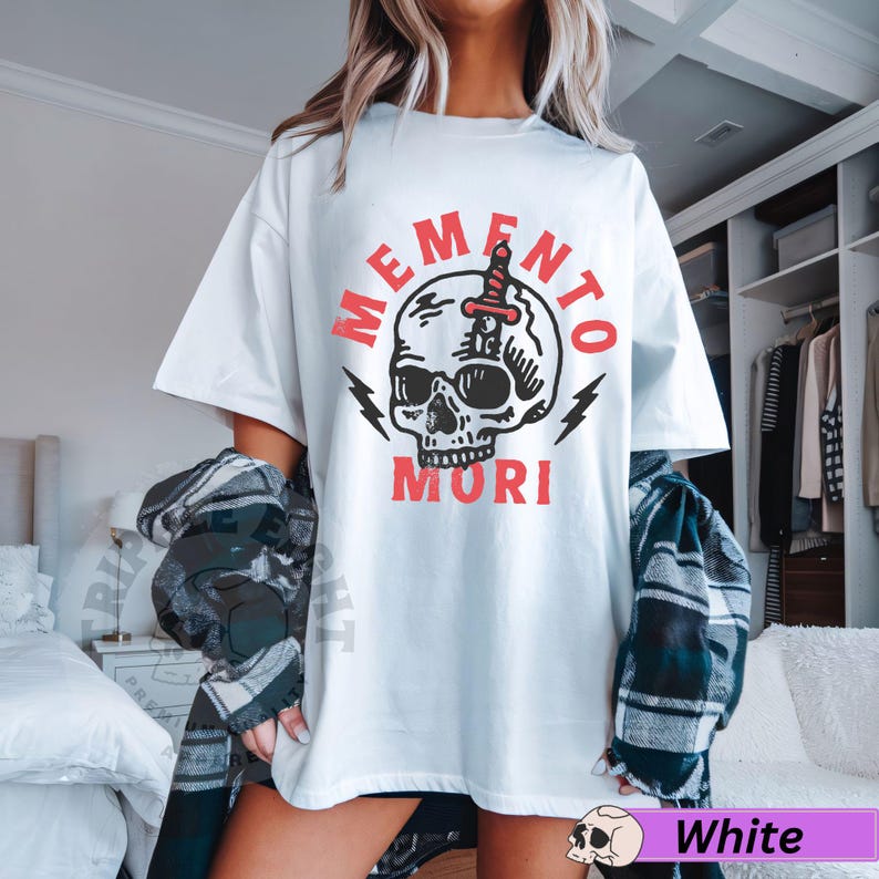 Puede incluir: Camiseta blanca con un gr&aacute;fico de calavera y el texto "Memento Mori" en rojo.