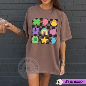 May include: Espresso-colored t-shirt with a colorful graphic of lucky charms on a checkered background. The design includes a four-leaf clover, horseshoe, rainbow, heart, and other symbols. The shirt has short sleeves and a crew neck.