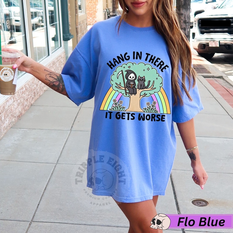 May include: A light blue t-shirt with a graphic of a cartoon grim reaper sitting on a tree with a cat. The text on the shirt reads "Hang in there it gets worse".