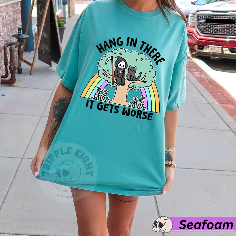 May include: A turquoise blue t-shirt with a graphic of a black cat and the Grim Reaper sitting on a tree with a rainbow behind them. The text on the shirt reads "Hang in there it gets worse".