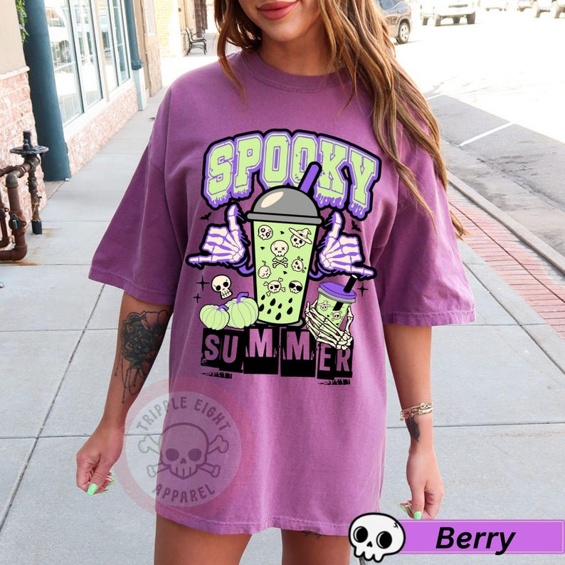 Puede incluir: Camiseta de color bayas con un gr&aacute;fico que dice "Spooky Summer". El dise&ntilde;o presenta una bebida boba verde con calaveras y huesos cruzados, manos de esqueleto y calabazas. La camiseta tiene mangas cortas y un corte holgado.