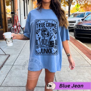 Puede incluir: Una camiseta azul con un gr&aacute;fico negro de un esqueleto sosteniendo una lupa y una copa de vino. El gr&aacute;fico est&aacute; en un marco con el texto "The True Crime Junkie".