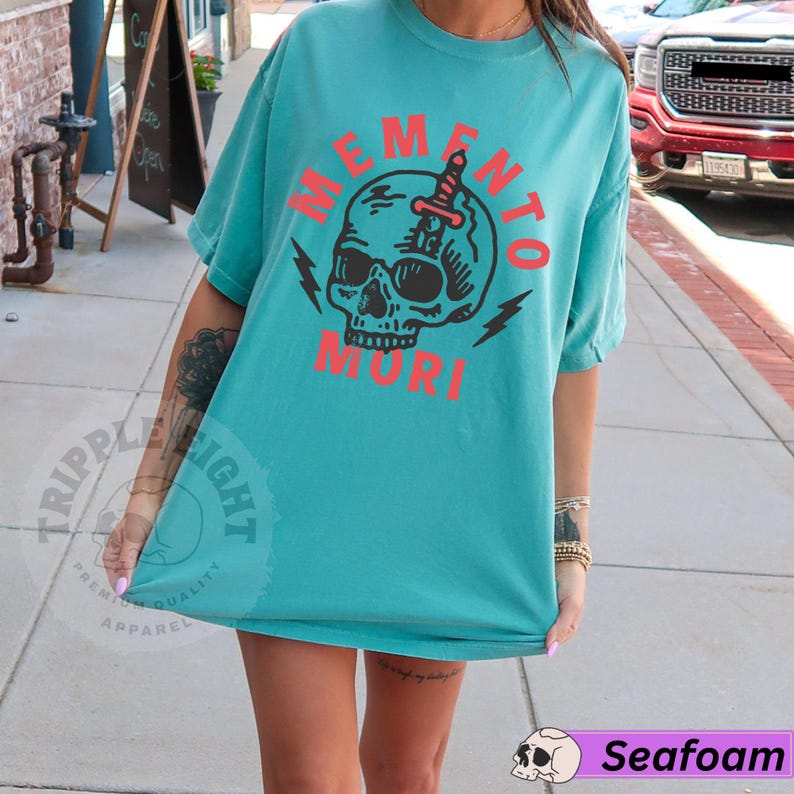 Puede incluir: Una camiseta azul turquesa con un gr&aacute;fico de calavera en blanco y negro y el texto "Memento Mori" en rojo.