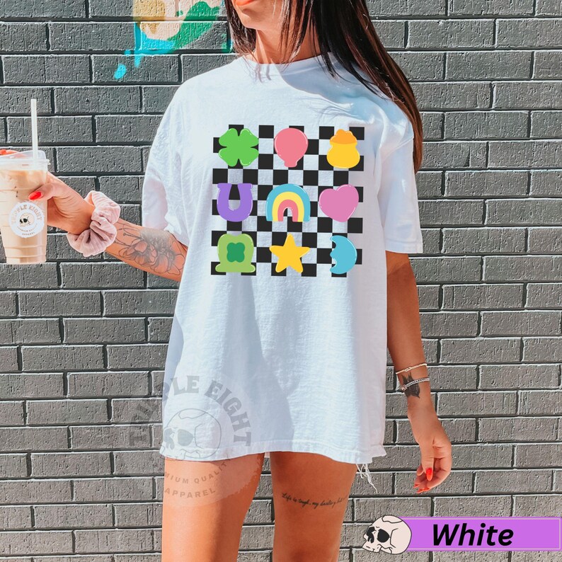 May include: White t-shirt with a colorful checkerboard design featuring lucky charms. The design includes a green four-leaf clover, pink balloon, yellow pot of gold, purple horseshoe, rainbow, pink heart, green shamrock, yellow star, and blue crescent moon.