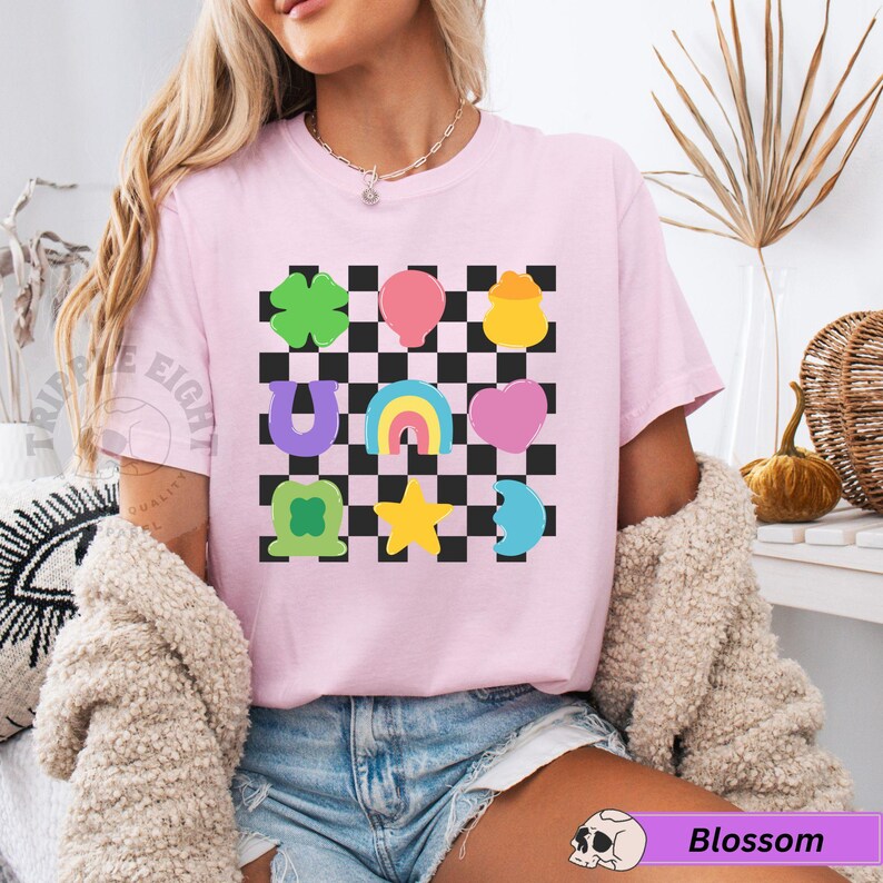 May include: A light pink t-shirt featuring a black and white checkered pattern with colorful graphic icons. The icons include a four-leaf clover, balloon, pot of gold, horseshoe, rainbow, heart, and star. The text "TRIPPLE EIGHT" is visible on the left side.