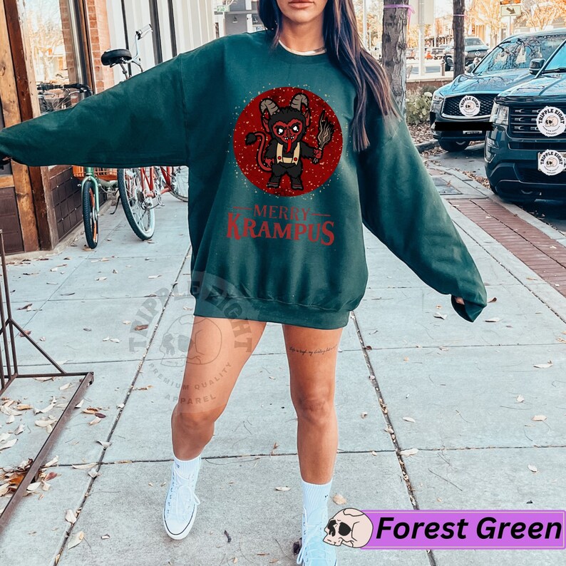 May include: A forest green sweatshirt with a red and black illustration of a Krampus figure holding a torch. The text "Merry Krampus" is printed in red below the illustration.