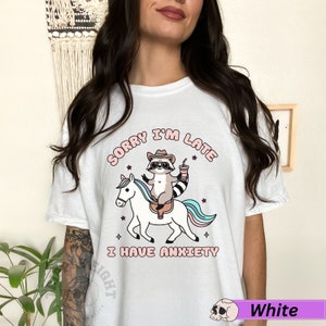 May include: White t-shirt with a graphic of a raccoon wearing a cowboy hat riding a unicorn. The text "Sorry I'm Late I Have Anxiety" is printed on the shirt.