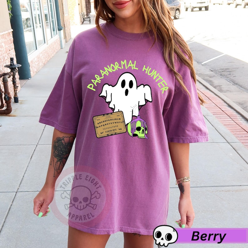 May include: Berry-colored t-shirt with a graphic that reads "PARANORMAL HUNTER" above a white ghost, a Ouija board, and a green skull. The shirt has short sleeves and a relaxed fit. The Triple Eight Apparel logo is visible.