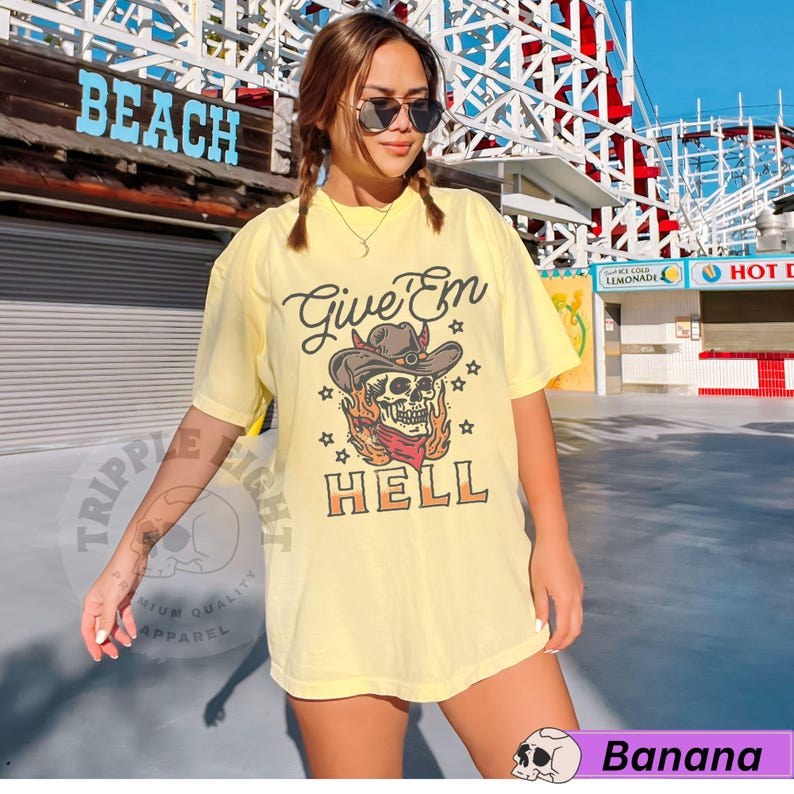 May include: Yellow t-shirt featuring a skull wearing a cowboy hat and bandana, with flames behind it. The text "Give 'Em Hell" is printed on the shirt.