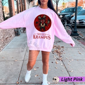 May include: A light pink sweatshirt with a red circle graphic of a cartoon devil holding a torch. The text "Merry Krampus" is printed below the graphic.