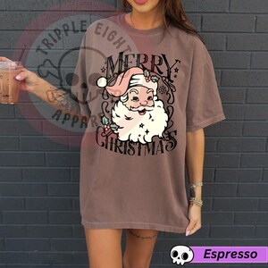 May include: Espresso-colored t-shirt featuring a vintage-style Santa Claus graphic with the words "MERRY CHRISTMAS". The Santa Claus illustration includes a pink hat, white beard, and holly accents. The shirt has a relaxed fit.