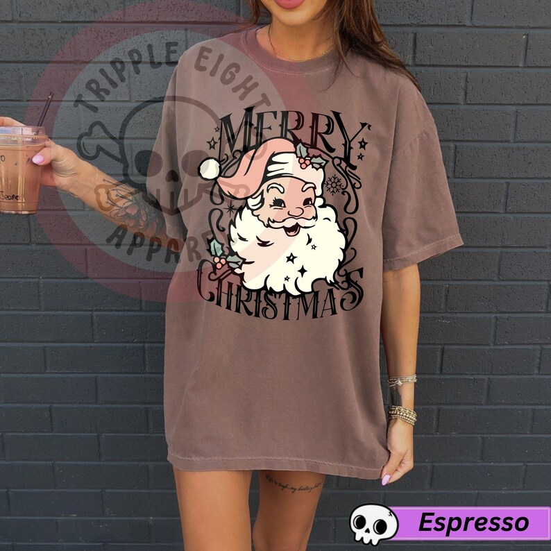 May include: Espresso-colored t-shirt featuring a vintage-style Santa Claus graphic with the words "MERRY CHRISTMAS". The Santa Claus illustration includes a pink hat, white beard, and holly accents. The shirt has a relaxed fit.