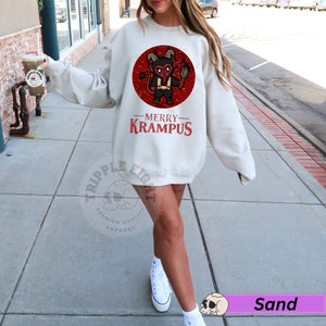 May include: A white sweatshirt with a red circle graphic featuring a cartoon Krampus holding a torch. The text "Merry Krampus" is printed below the graphic.