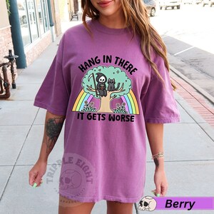 May include: Purple t-shirt with a graphic of a skeleton sitting on a tree with a cat. The graphic has a rainbow behind it and the text "Hang in there it gets worse".