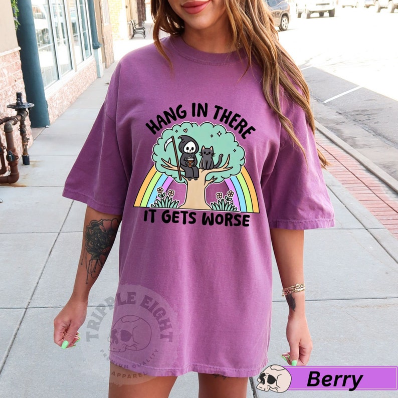 May include: Purple t-shirt with a graphic of a skeleton sitting on a tree with a cat. The graphic has a rainbow behind it and the text "Hang in there it gets worse".