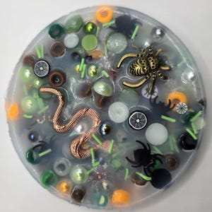 May include: A round, resin coaster with a Halloween theme. It features a copper-colored snake, a gold-colored spider, and several black spiders. The coaster is filled with various beads in shades of green, orange, and white.